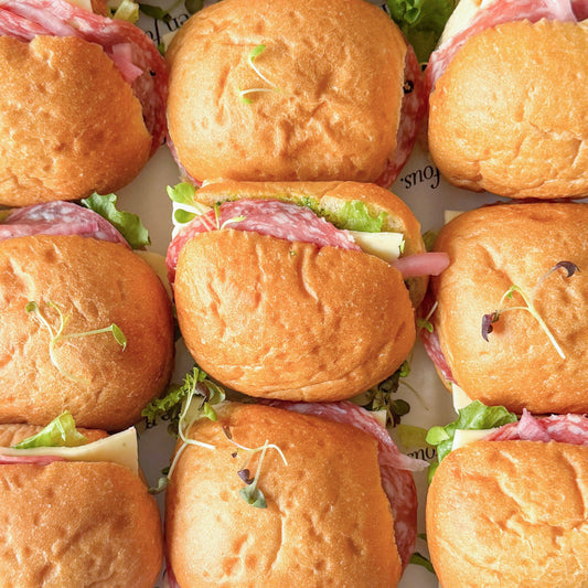 Overhead close-up of multiple slider sandwiches arranged in a grid, with golden-brown buns, sliced salami, white cheese, lettuce, and a few microgreens on top.