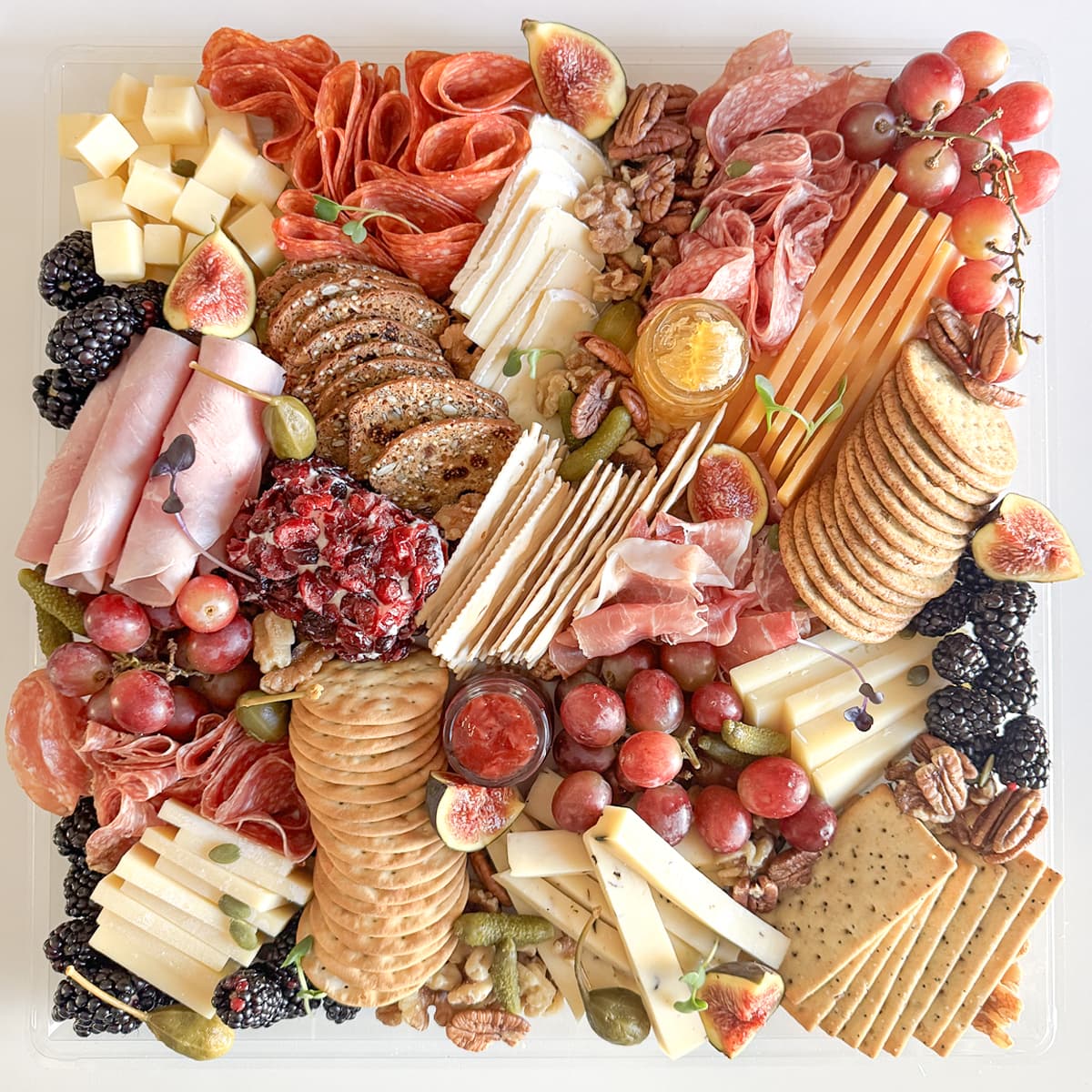 A top-down view of a large charcuterie board arranged in a clear square tray, featuring sliced cheeses, cured meats, crackers, grapes, figs, blackberries, nuts, and dried fruit on a white background.