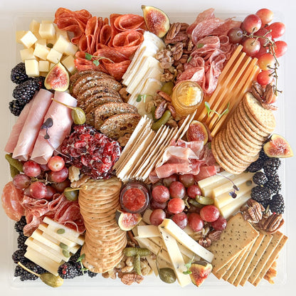 A top-down view of a large charcuterie board arranged in a clear square tray, featuring sliced cheeses, cured meats, crackers, grapes, figs, blackberries, nuts, and dried fruit on a white background.