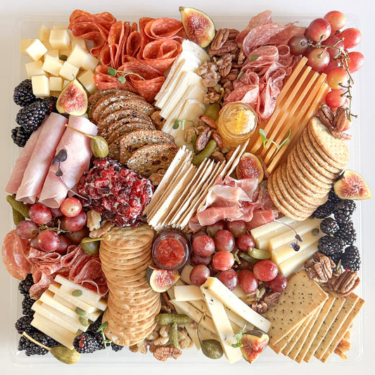 A top-down view of a large charcuterie board arranged in a clear square tray, featuring sliced cheeses, cured meats, crackers, grapes, figs, blackberries, nuts, and dried fruit on a white background.