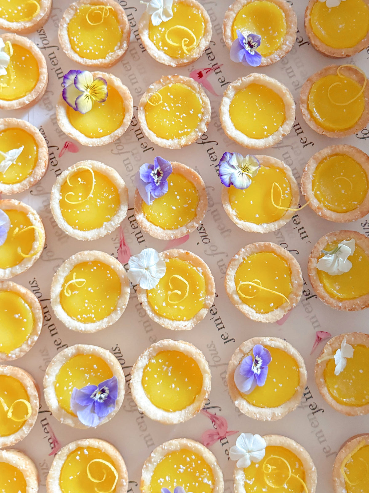A top-down view of multiple small lemon tartlets filled with bright yellow custard, arranged in rows on a light background with decorative script and edible flowers.