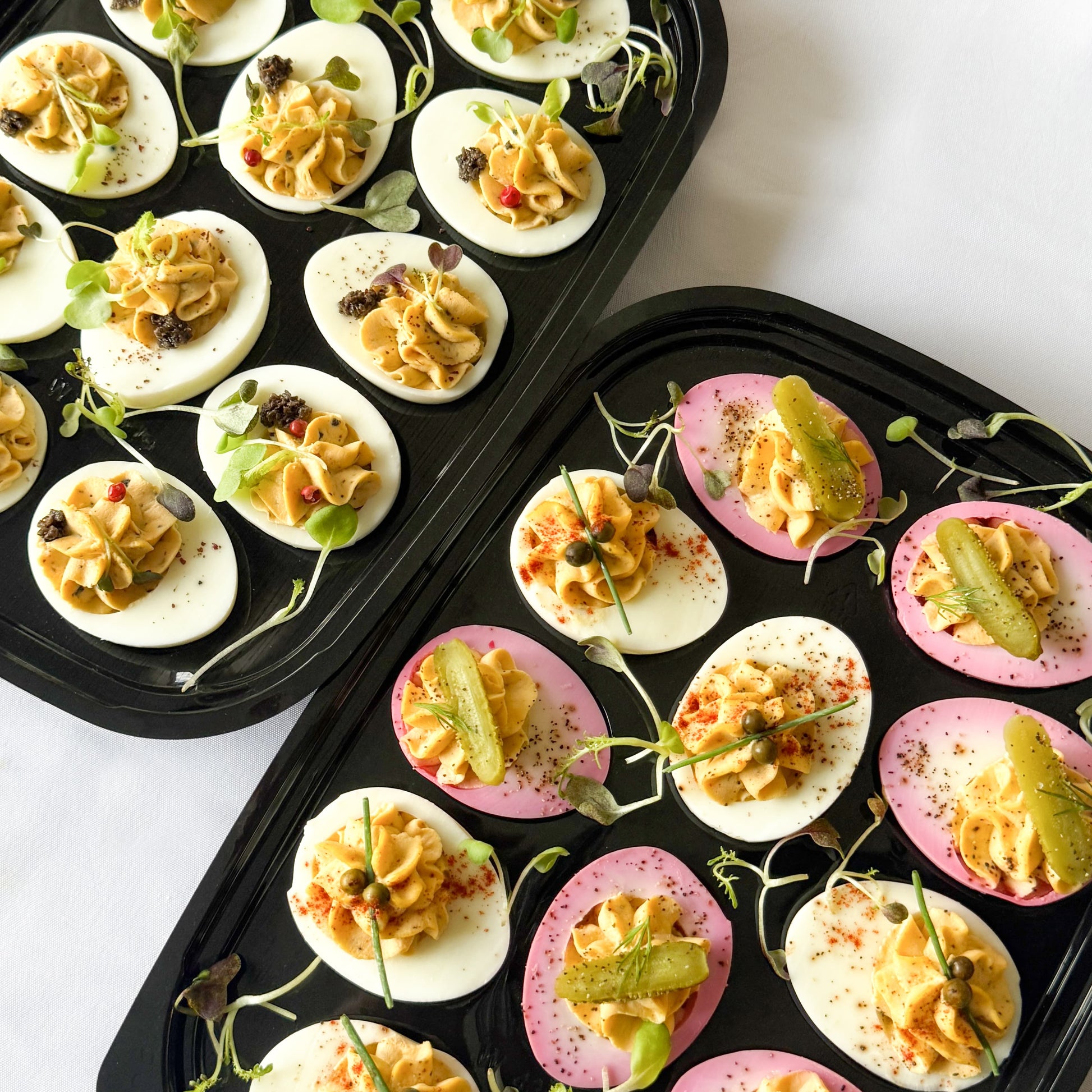 Two ready to serve trays filled with assorted truffle and pickled deviled eggs garnished with herbs and pickles on a white background.