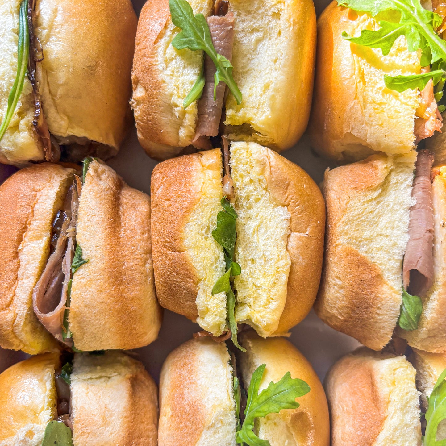Brioche sliders with roast beef, arugula and aioli arranged in rows in a box.