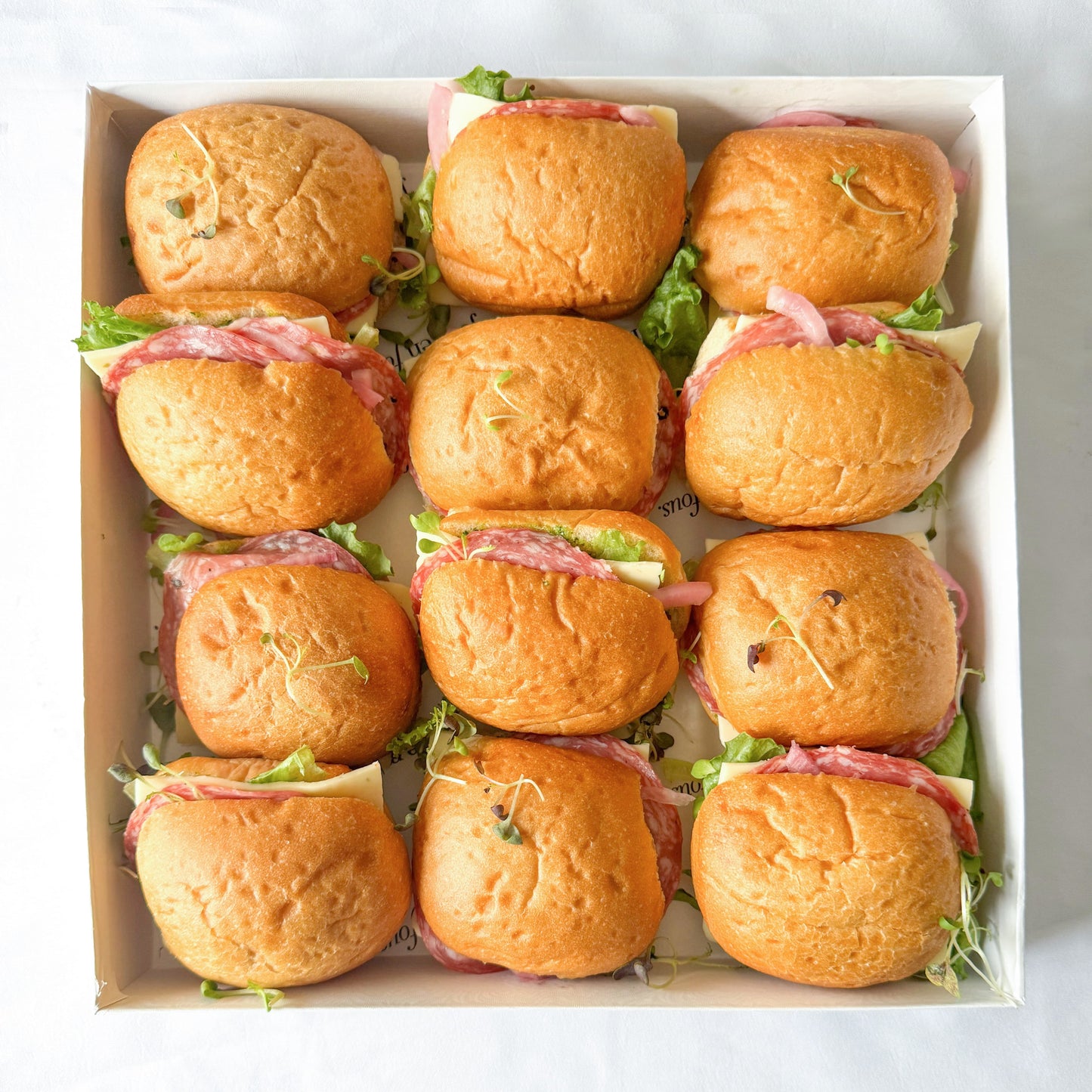 Box of mini sandwiches with salami, lettuce, cheese on a white background.