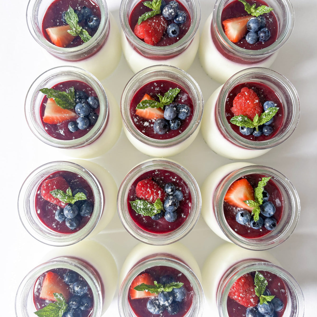 Twelve small glass jars filled with white panna cotta, topped with dark berry coulis, fresh strawberries, blueberries, and mint leaves, arranged in a neat grid on a white background.