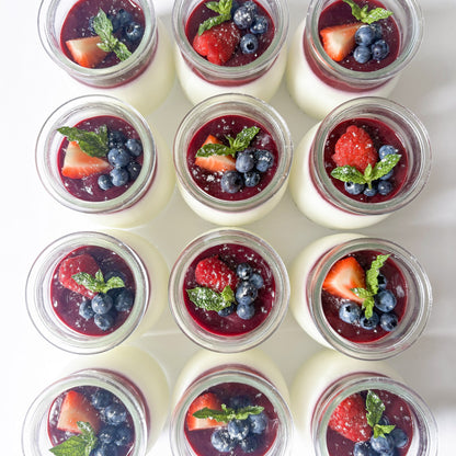 Twelve small glass jars filled with white panna cotta, topped with dark berry coulis, fresh strawberries, blueberries, and mint leaves, arranged in a neat grid on a white background.