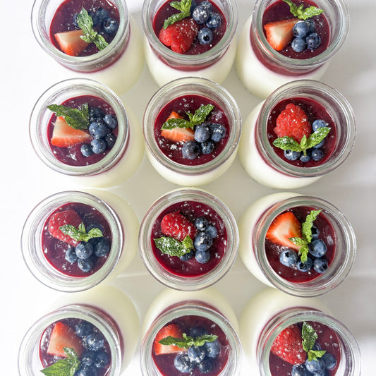 Twelve small glass jars filled with white panna cotta, topped with dark berry coulis, fresh strawberries, blueberries, and mint leaves, arranged in a neat grid on a white background.