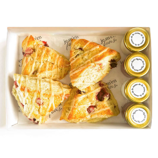 Box of triangle orange cranberry scones with jars of jam and clotted cream on a white background.
