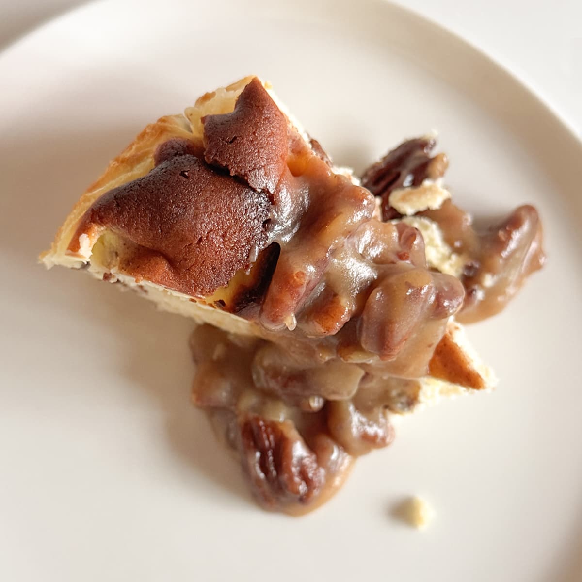 A close-up shot of a slice of cheesecake topped with gooey pecan pie filling on a white plate.