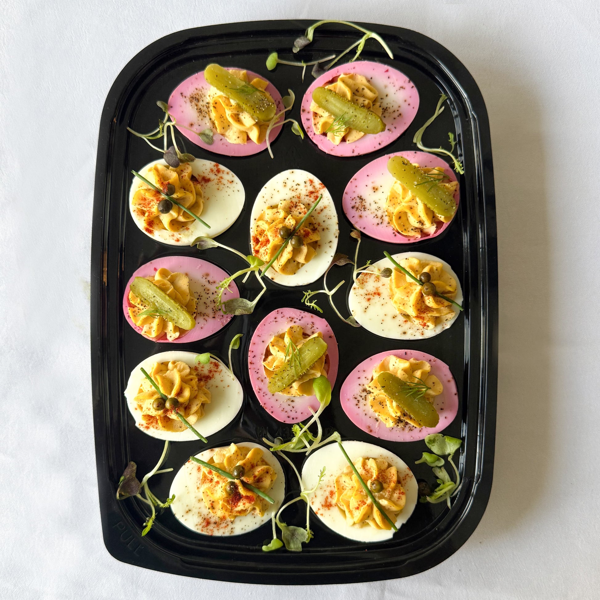 Ready to serve tray filled with classic and pink pickled deviled eggs garnished with microgreens and mini pickles on a white background.