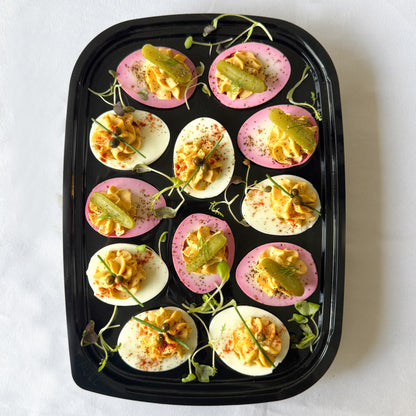Ready to serve tray filled with classic and pink pickled deviled eggs garnished with microgreens and mini pickles on a white background.