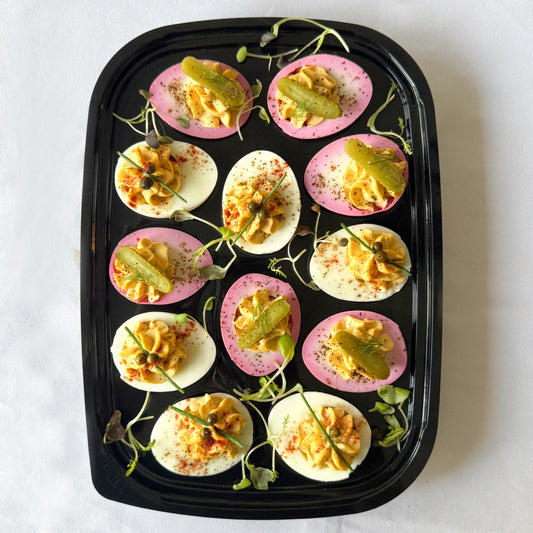 Ready to serve tray filled with classic and pink pickled deviled eggs garnished with microgreens and mini pickles on a white background.