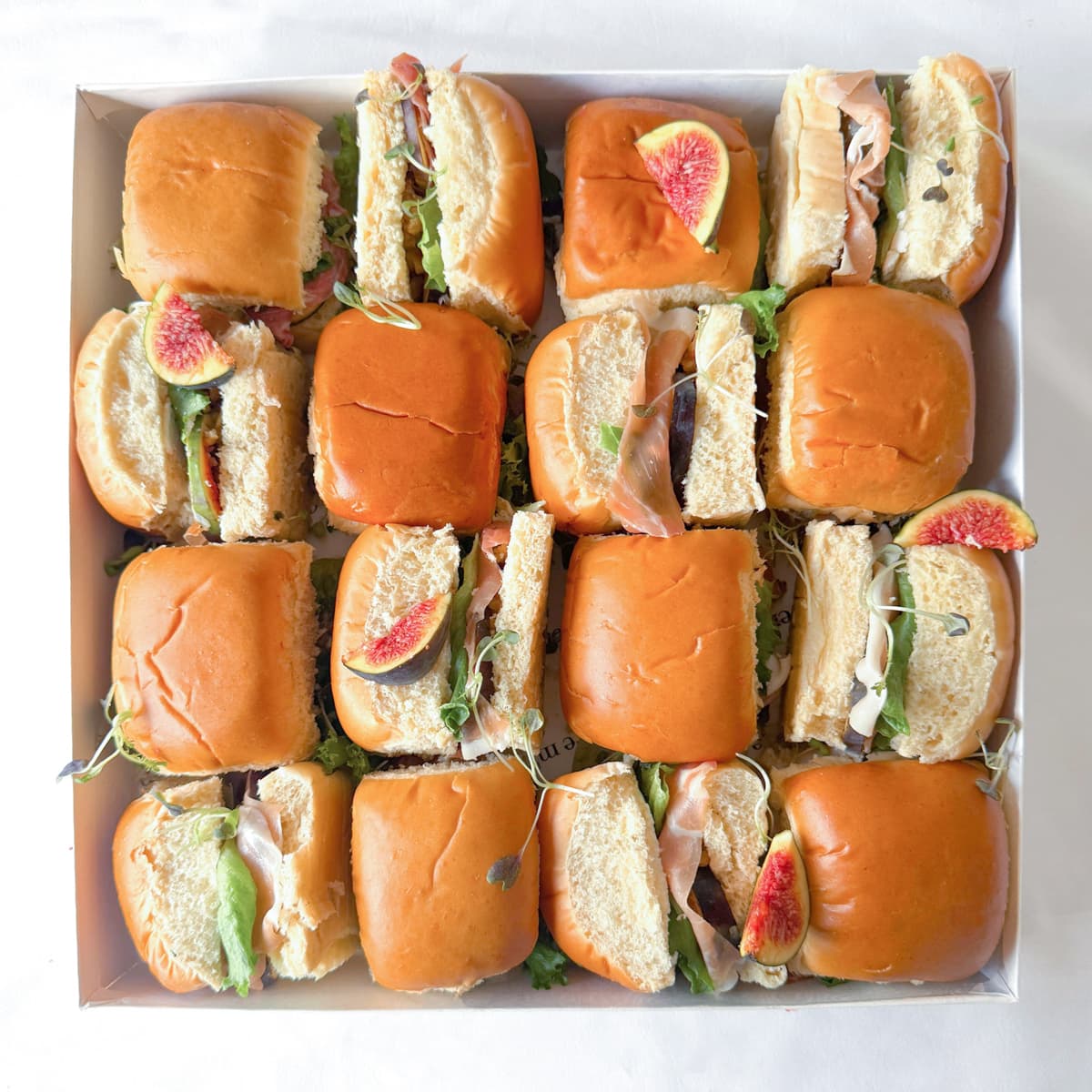 Box of mini brioche sandwiches with proscuitto, figs and lettuce on a white background.
