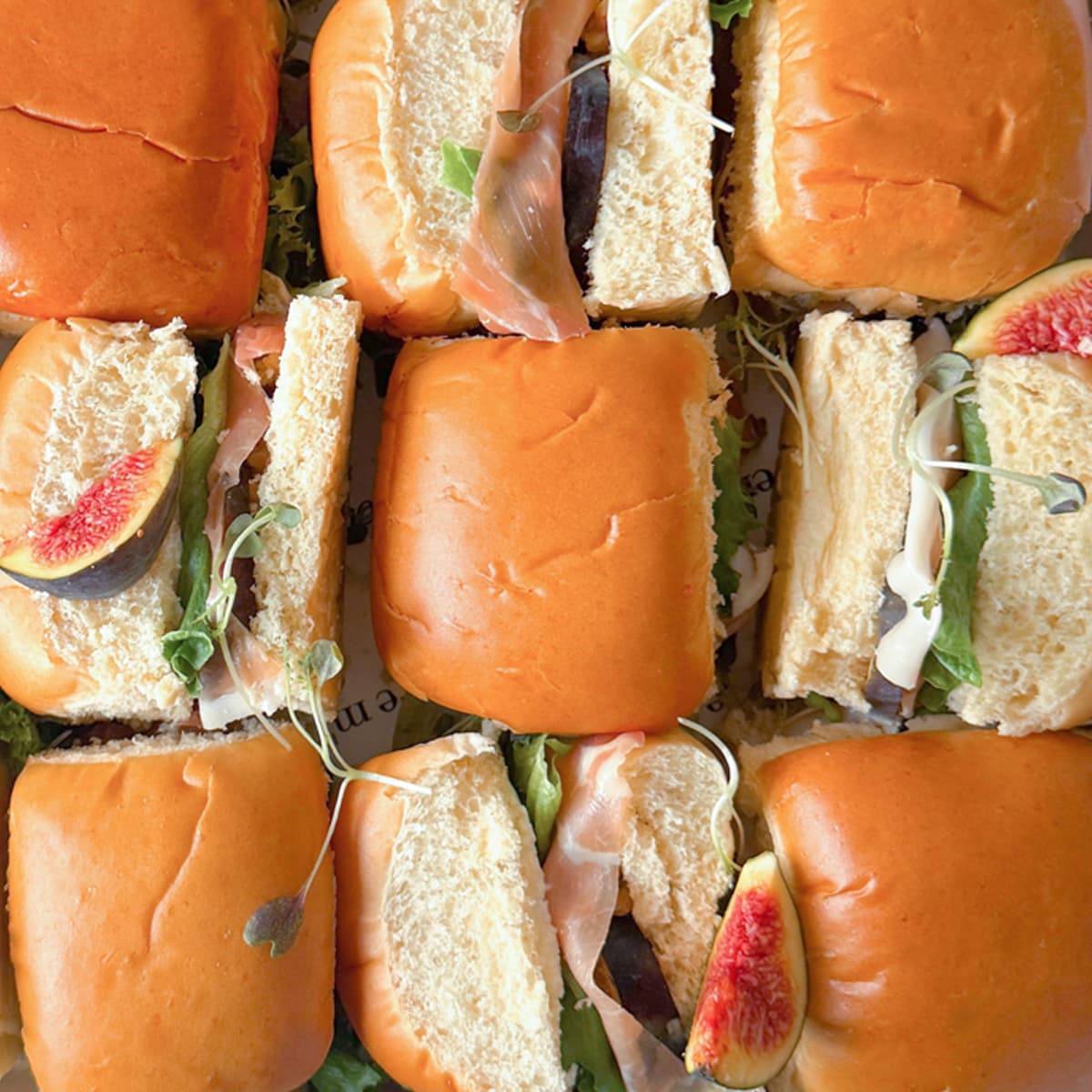 Box of mini brioche sandwiches with proscuitto, figs and lettuce on a white background.