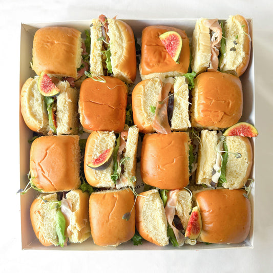 Box of mini brioche sandwiches with proscuitto, figs and lettuce on a white background.