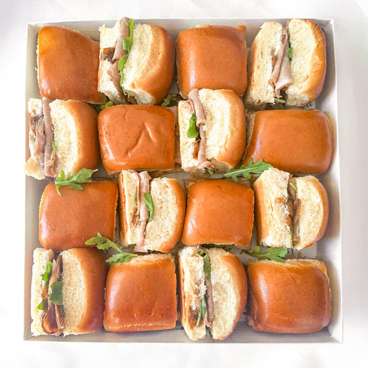 A top-down view of a boxed set of 16 mini sliders arranged in a white tray. The sliders feature glossy golden-brown buns, sliced roast beef, caramelized onions and fresh green arugula leaves.