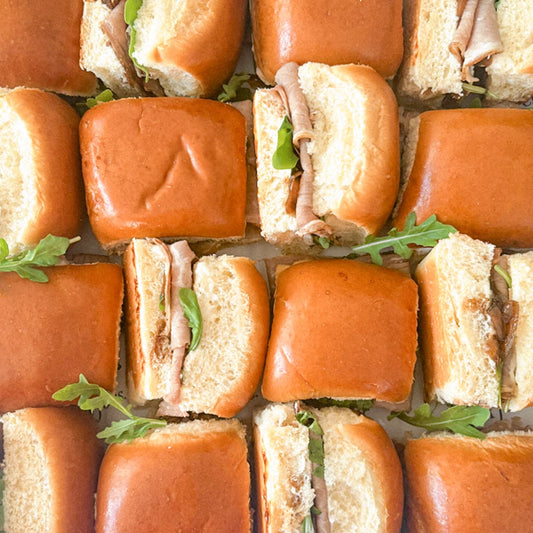 Overhead view of multiple mini slider sandwiches arranged in a grid, with glossy golden-brown buns, sliced roast beef, caramelized onions and green arugula leaves.