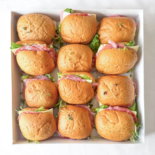 A top-down view of a white cardboard tray holding 12 mini sliders filled with italian salami ham, cheese, and greens.