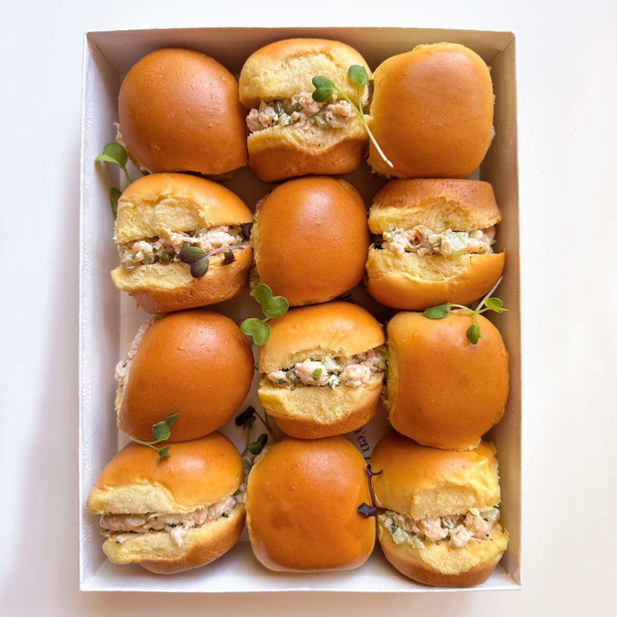 A top-down view of a white cardboard tray holding twelve mini sliders filled with shrimp salad, garnished with small green microgreens, on a white background.