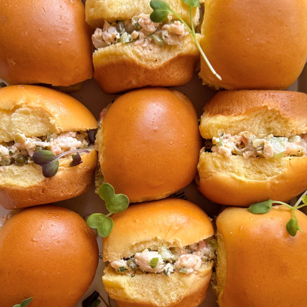 A close-up top-down view of eight golden-brown slider rolls filled with a creamy shrimp salad, garnished with small green microgreens.