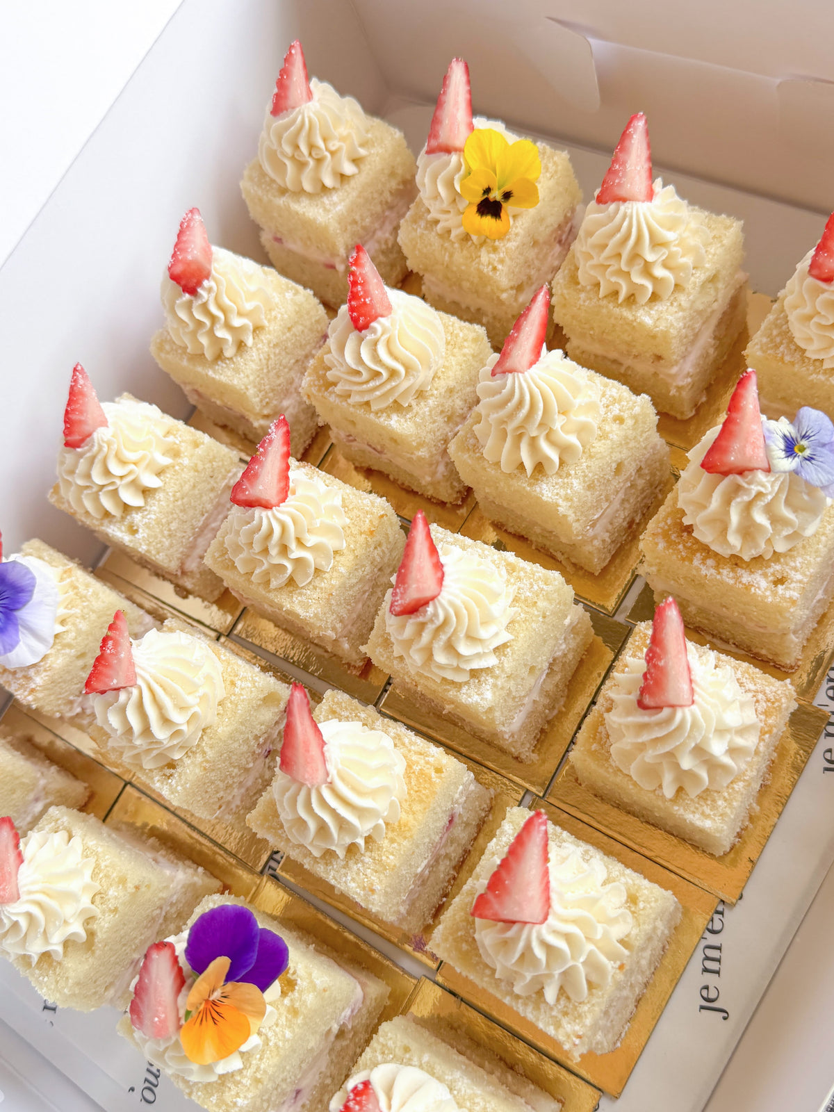 A box of small square sponge cake slices topped with piped cream, sliced strawberries, and edible flowers, arranged in a clear tray inside a white box.