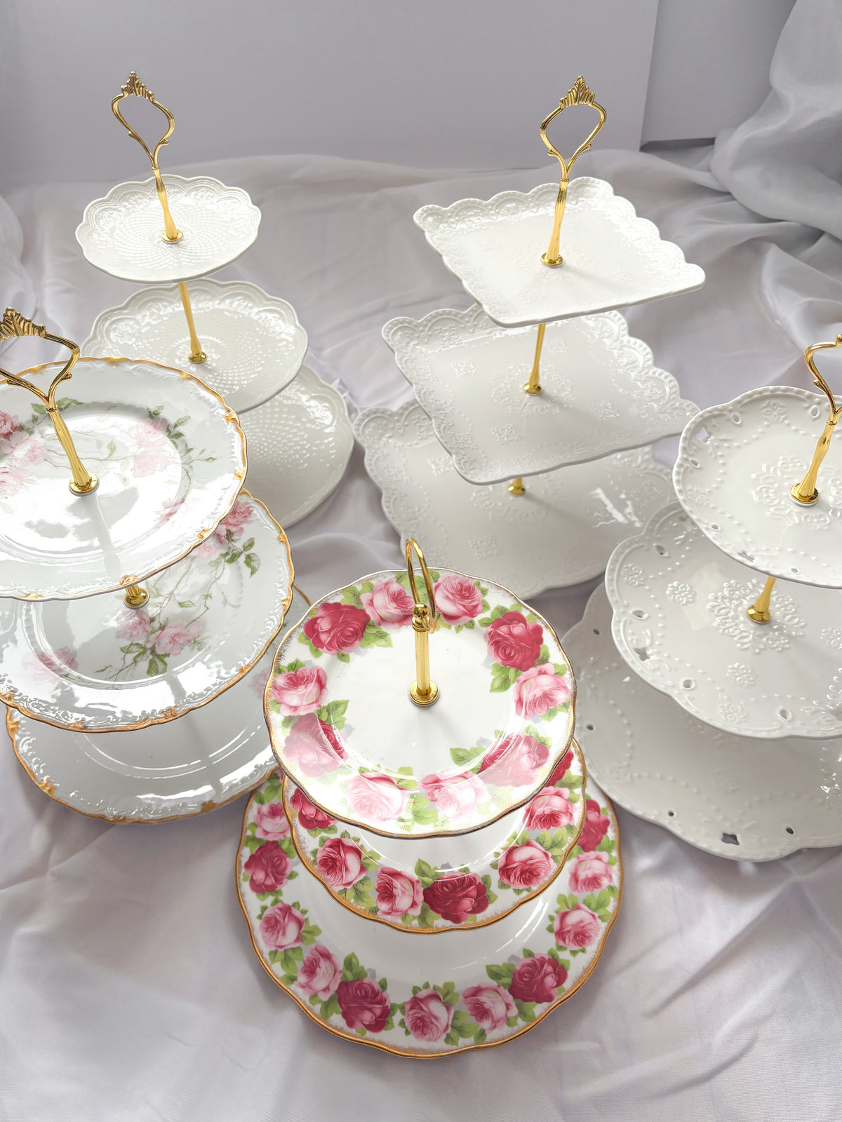 A collection of white ceramic three-tiered serving stands decorated with pink rose patterns and gold accents, arranged on draped white fabric.