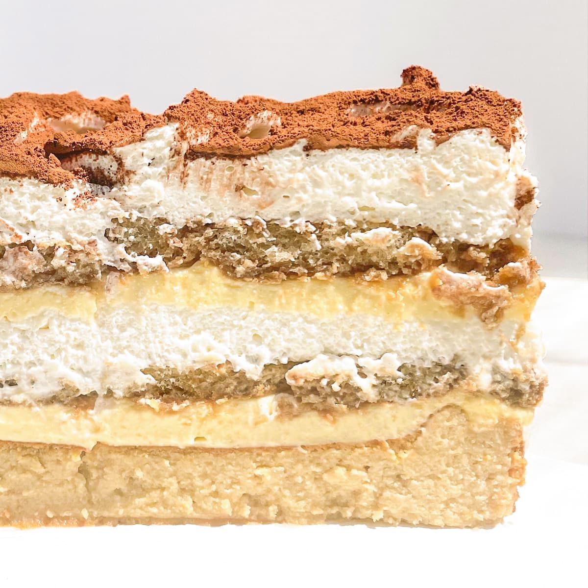 Tiramisu cake cut in half, a basque cheesecake base then topped with several layers of whipped cream, coffee lady fingers and cocoa powder.