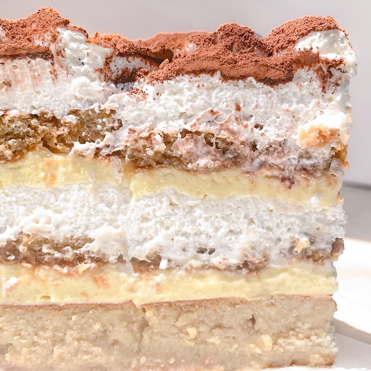 A close-up side view of a layered tiramisu cake, showing alternating layers of whipped cream, yellow pastry cream, soaked coffee lady finger cookies, and a dusting of cocoa powder on top.