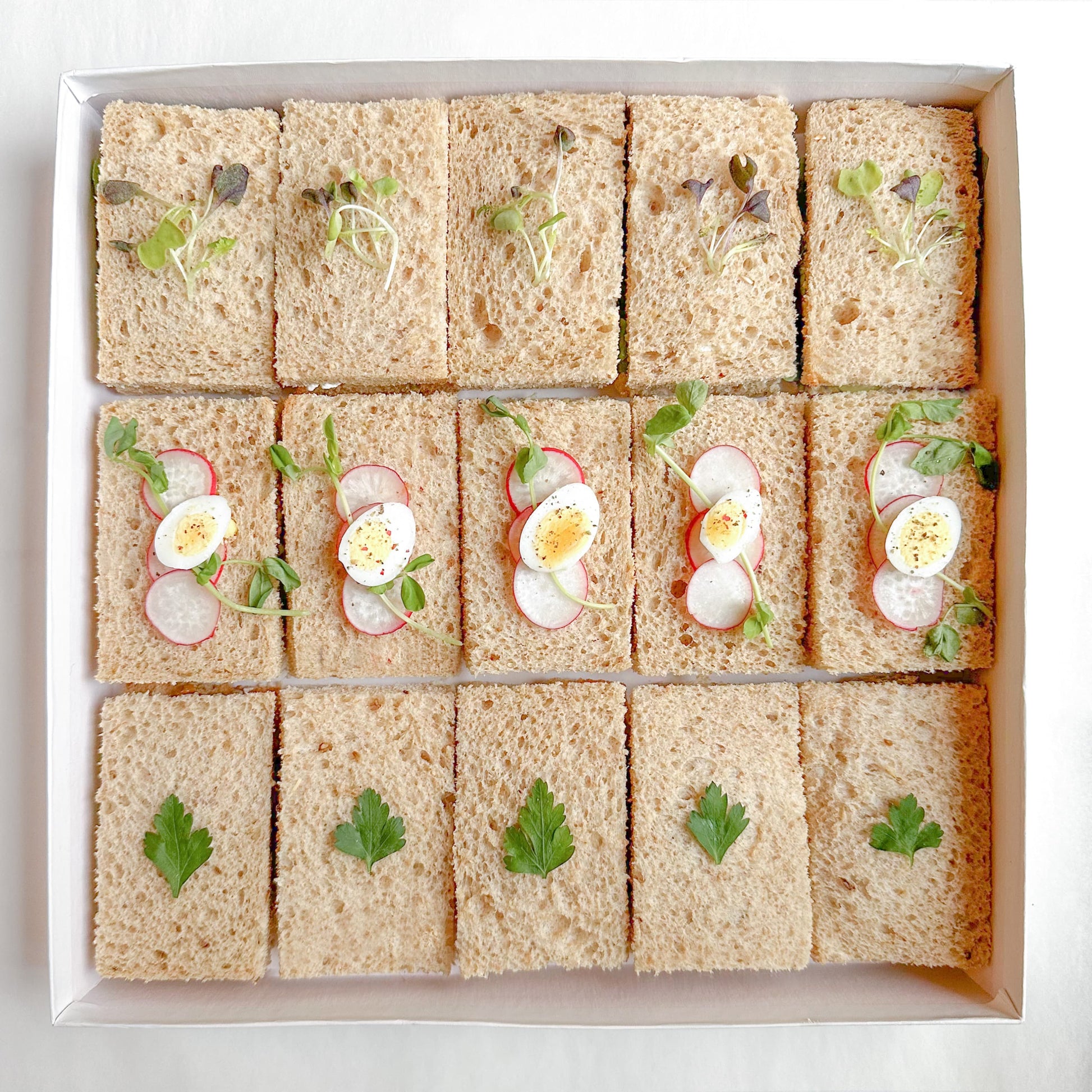 A box of prepared vegetarian mini tea sandwiches with various fillings such as egg salad, roasted pepper, avocado and hummus, and green goddess on brown bread.