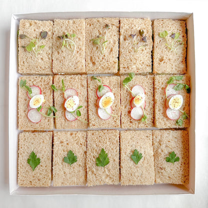 A box of prepared vegetarian mini tea sandwiches with various fillings such as egg salad, roasted pepper, avocado and hummus, and green goddess on brown bread.