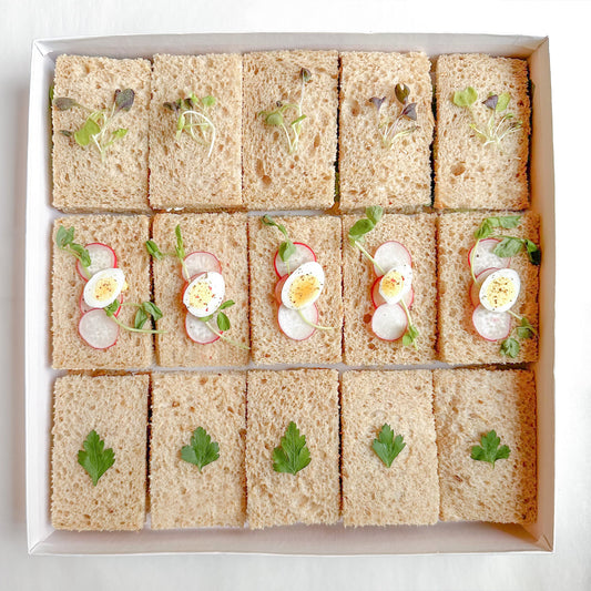 A box of prepared vegetarian mini tea sandwiches with various fillings such as egg salad, roasted pepper, avocado and hummus, and green goddess on brown bread.