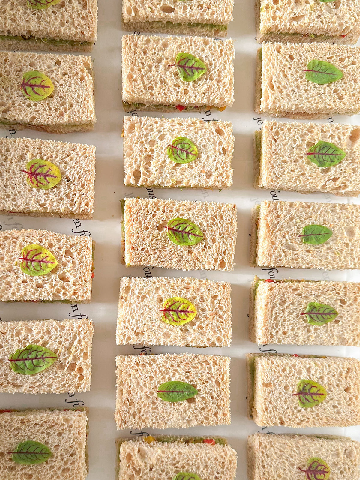 A top-down view of multiple mini sandwiches arranged in rows on a white surface, each topped with a small sorrel leaf.